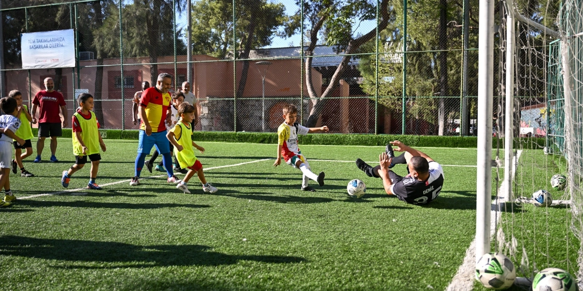 Açık hava halı sahasında futbol antrenmanı yapan çocuklar ve onlara eşlik eden yetişkinler. Bir çocuk topa şut çekerken, kaleci pozisyonundaki yetişkin topu kurtarmaya çalışıyor. Saha kenarında antrenörler ve diğer çocuklar izliyor, arka planda ise üzerinde 