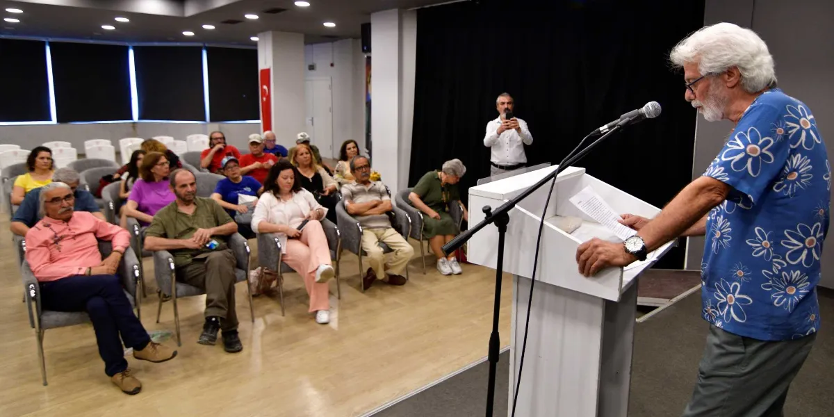 Beyaz saçlı ve sakallı, mavi çiçek desenli bir gömlek giyen bir adam, kürsüde elindeki kağıttan şiir okuyor. Karşısında, kapalı bir salonda gri koltuklarda oturan ve onu dikkatle dinleyen yaklaşık on beş kişilik bir izleyici grubu bulunuyor. Arka planda ayakta duran bir kişi ise cep telefonuyla çekim yapıyor.