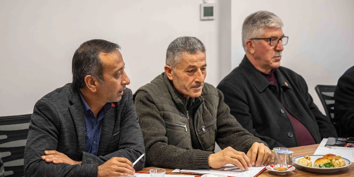 Toplantı masasında yan yana oturan üç erkek, önlerindeki evrakları inceliyor. En soldaki kişi koyu renkli ceket ve desenli gömlek giymiş, elinde bir kalem tutarak yanındaki kişiye bakıyor. Ortadaki kişi gri saçlı, bıyıklı, haki renkli şişme yelek ve kazak giymiş durumda. En sağdaki kişi ise gözlüklü, gri saçlı, siyah ceket ve bordo kazak giymiş şekilde karşıya bakıyor. Masanın üzerinde ince belli çay bardağı ve küçük atıştırmalıkların bulunduğu bir tabak yer alıyor.