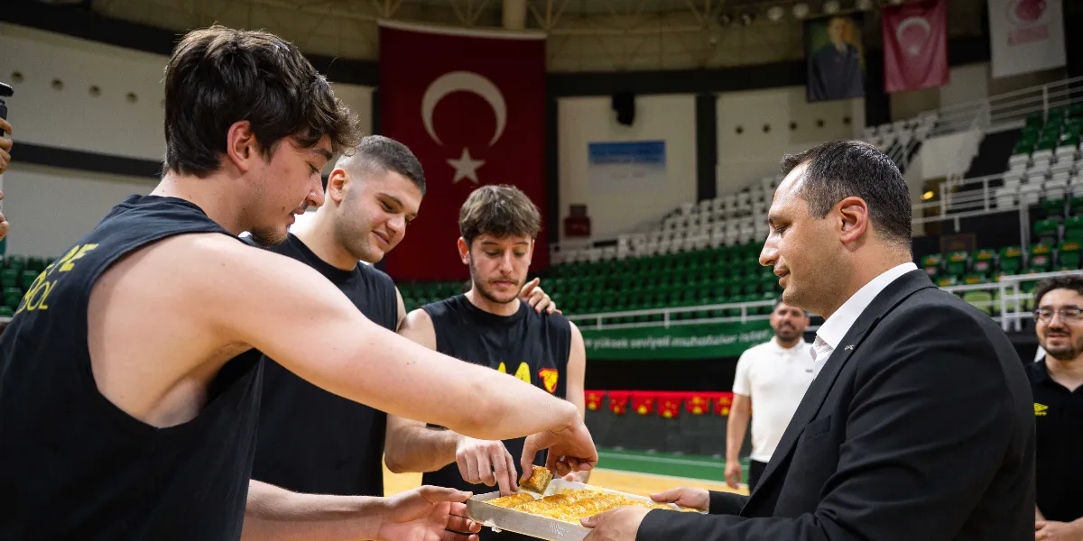 Siyah takım forması giyen üç genç basketbolcu, kapalı bir spor salonunda kendilerini ziyaret eden ve elinde baklava tepsisi tutan takım elbiseli bir yetkiliye gülümseyerek bakıyor. Basketbolculardan biri tepsiden bir dilim baklava alırken, arka planda Türk bayrağı ve boş tribünler görülüyor.
