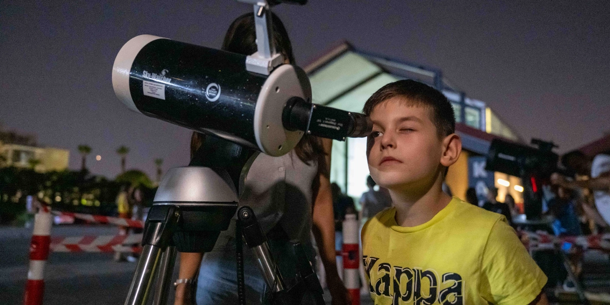 Gece vakti düzenlenen bir gökyüzü gözlem etkinliğinde, sarı tişörtlü küçük bir erkek çocuk Sky-Watcher marka bir teleskobun merceğine bakıyor. Çocuğun arkasında bir yetişkin duruyor ve arka planda aydınlatılmış bir yapı ile başka gözlemciler seçiliyor.