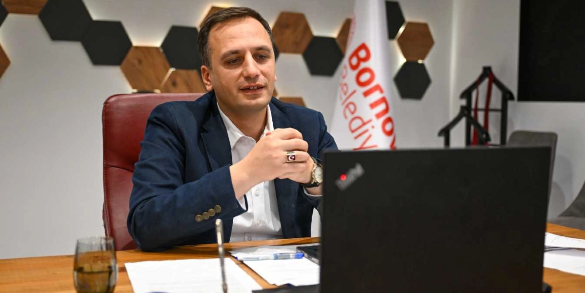 Koyu renk ceket ve beyaz gömlek giymiş, parmağında Türk bayrağı işlemeli bir yüzük bulunan bir erkek, ofis ortamında masasında oturarak dizüstü bilgisayar ekranına bakıyor. Arka planda Bornova Belediyesi yazılı bir bayrak ve duvarda altıgen dekoratif paneller yer alıyor.