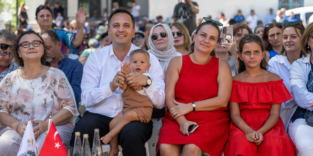 Açık havada düzenlenen bir etkinlikte, ön sırada yan yana oturan bir grup insan. Merkezde beyaz gömlekli bir erkek, kucağında küçük bir bebekle oturuyor; yanında kırmızı kolsuz bir elbise giyen bir kadın ve onun yanında yine kırmızı, omuzları açık bir elbise giyen genç bir kız bulunuyor. Grubun önündeki masada Türk bayrağı ve cam şişeler yer alıyor. Arka planda ise etkinliği izleyen kalabalık bir grup insan görülüyor.