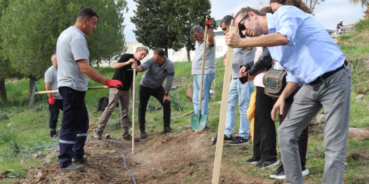 Açık havada, yeşil bir alanda bir grup insan toprak zeminde çalışma yapıyor. Ön planda açık mavi gömlekli bir adam, elinde tuttuğu ahşap bir çubuğu toprağa yerleştirirken görülüyor. Yanında kırmızı eldivenli bir çalışan, elinde bir tırmıkla duruyor. Diğer kişiler ise ellerinde küreklerle toprağı kazıyor ve yerleştirilen çubukları kontrol ediyor. Arka planda ağaçlar ve hafif eğimli bir arazi bulunuyor.