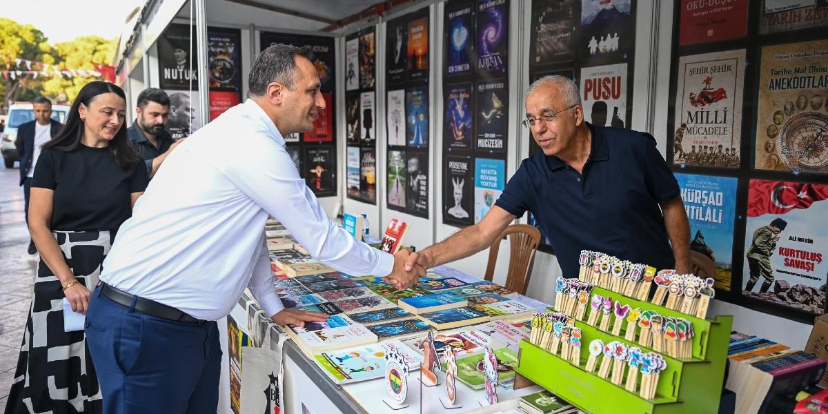Açık havada kurulan bir kitap fuarı standında, beyaz gömlekli bir erkek ile lacivert polo yaka tişörtlü, gözlüklü bir erkek el sıkışıyor. Standın üzerinde çeşitli kitaplar ve üzerinde farklı figürler bulunan kitap ayraçları sergileniyor. Arka planda duvarda Nutuk, Milli Mücadele ve Kurtuluş Savaşı gibi temalara sahip kitap kapakları asılı. Sol tarafta siyah bluz ve desenli etek giymiş bir kadın, sağ tarafta ise sakallı başka bir erkek onları izliyor.