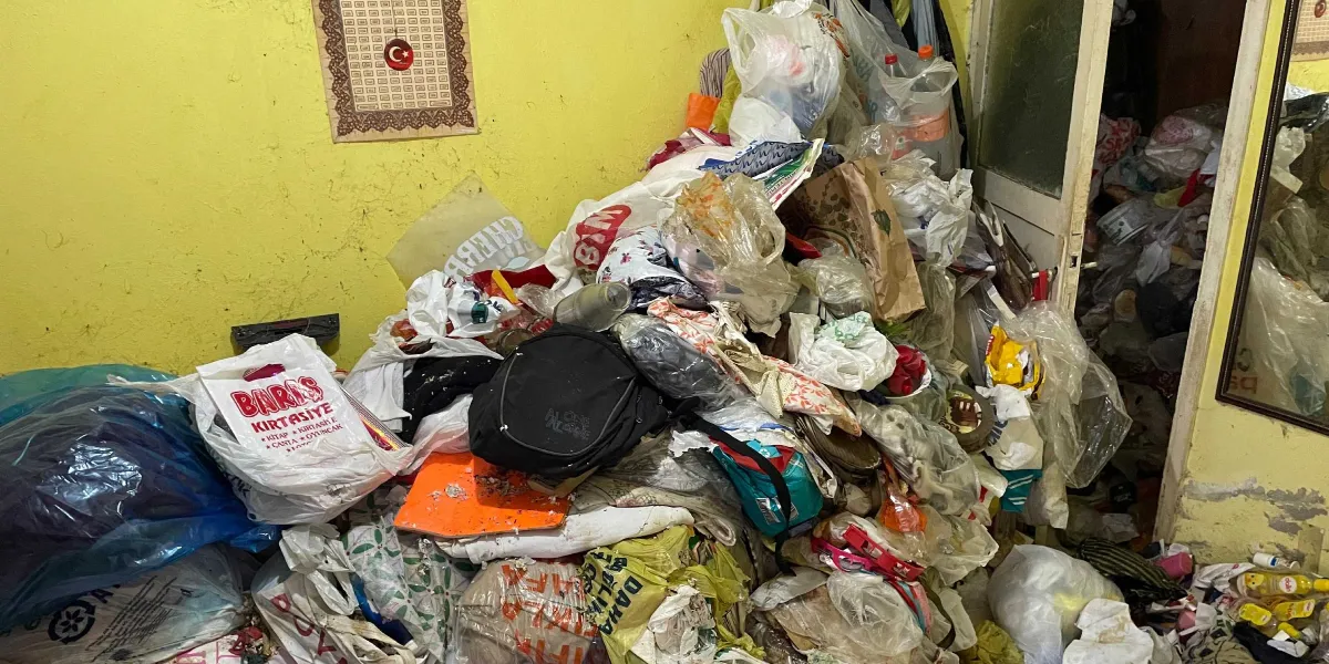 Sarı duvarlı bir odanın içinde, yerlere ve köşelere yığılmış çok miktarda çöp, poşet, eski eşya ve atık malzemelerden oluşan sağlıksız yaşam alanı. Duvarın bir kısmında asılı bir Türk bayrağı motifi bulunurken, odanın kapı eşiği ve zemin tamamen atıklarla kaplanmış durumdadır.