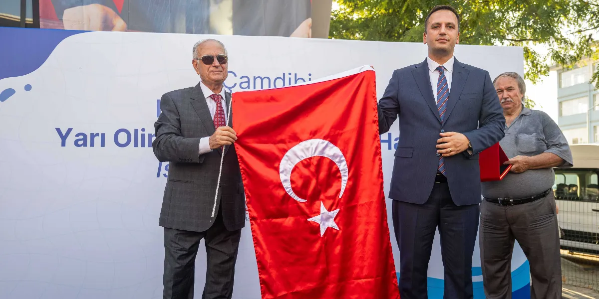 Bornova'da düzenlenen bir törende, takım elbiseli iki erkek yan yana durarak ellerinde tuttukları Türk bayrağını açıyor. Sol taraftaki kişi güneş gözlüğü takıyor, sağ taraftaki kişi ise lacivert takım elbise ve kravat giyiyor. Arka planda üzerinde 