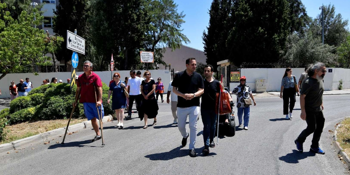 Açık havada, asfalt bir yolda yürüyen çeşitli yaş ve fiziksel özelliklere sahip bir grup insan. Grubun önünde koltuk değneği kullanan bir adam, yanında tekerlekli sandalye kullanan bir bireye eşlik eden bir kişi ve diğer katılımcılar yer alıyor. Arka planda ağaçlar, Bornova İlçe Tarım ve Orman Müdürlüğü tabelası ve beyaz bir duvar bulunuyor. Gün ışığında çekilmiş bu kare, engelli hakları konusunda farkındalık yaratmak amacıyla yapılan bir yürüyüşü yansıtıyor.
