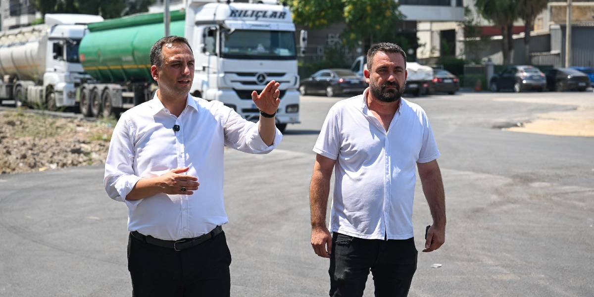 Beyaz gömlekli iki erkek, asfalt bir alanda yürürken bir şeyler konuşuyor. Soldaki kişi elini havaya kaldırarak bir yeri işaret ediyor, sağdaki kişi ise onu dinliyor. Arka planda üzerinde KILIÇLAR yazan beyaz bir tır ve yeşil bir tanker ile park halindeki otomobiller görülüyor.