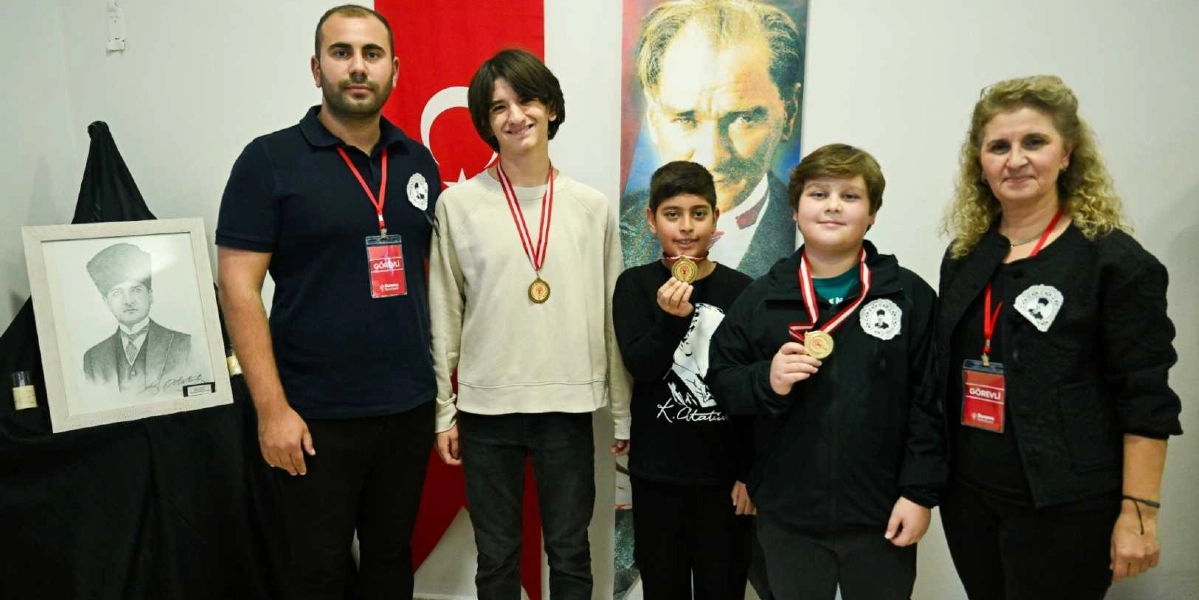 Arka planda Türk bayrağı ve Mustafa Kemal Atatürk'ün portresinin yer aldığı bir etkinlikte, boyunlarında madalyalarla yan yana duran iki yetişkin ve üç çocuk. Sol tarafta siyah bir örtü üzerinde Atatürk'ün çerçeveli bir karakalem çizimi bulunuyor. Yetişkinlerin üzerinde 