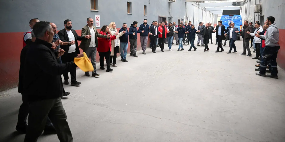 Belediye Başkanı Ömer Eşki ve bir grup işçi, gri duvarlı bir avluda el ele tutuşarak halay çekiyor. Ön planda elinde sarı bir mendil tutan bir kişi halayı yönlendirirken, arka planda üzerinde 