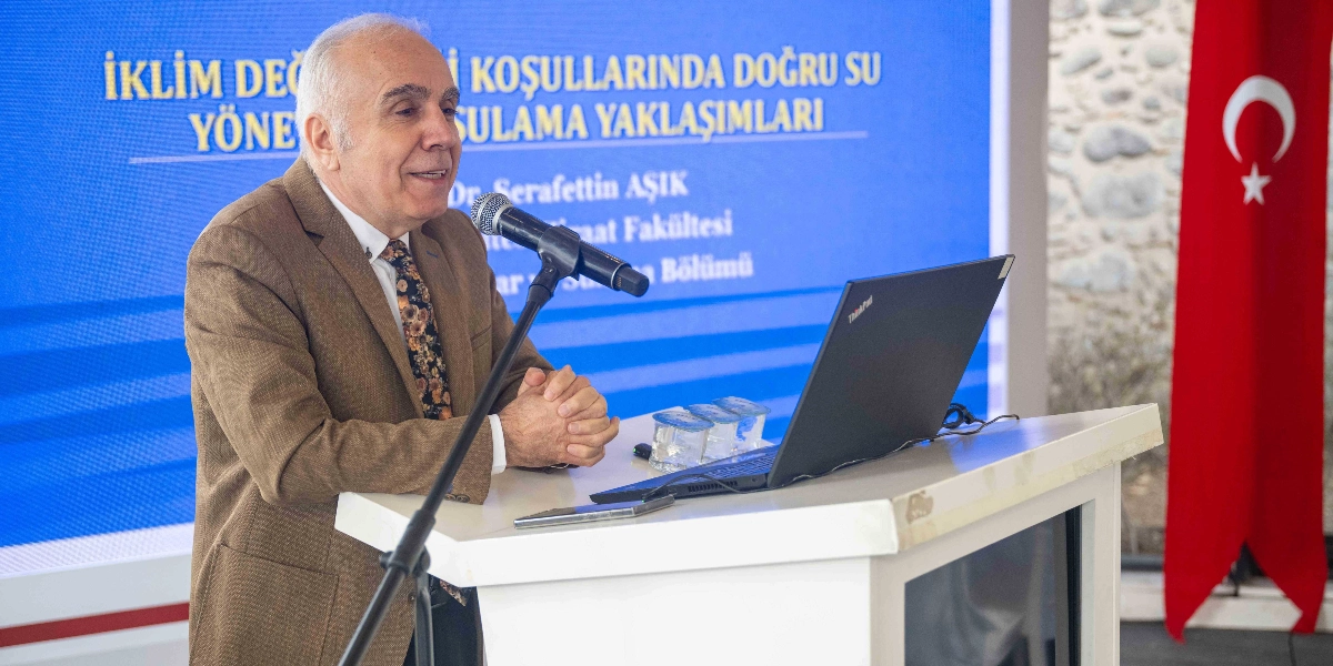 Kahverengi ceketli ve desenli kravatlı Dr. Şerafettin Aşık, kürsüde mikrofon başında konuşma yapıyor. Önünde açık bir dizüstü bilgisayar ve su bardakları bulunuyor. Arka planda 