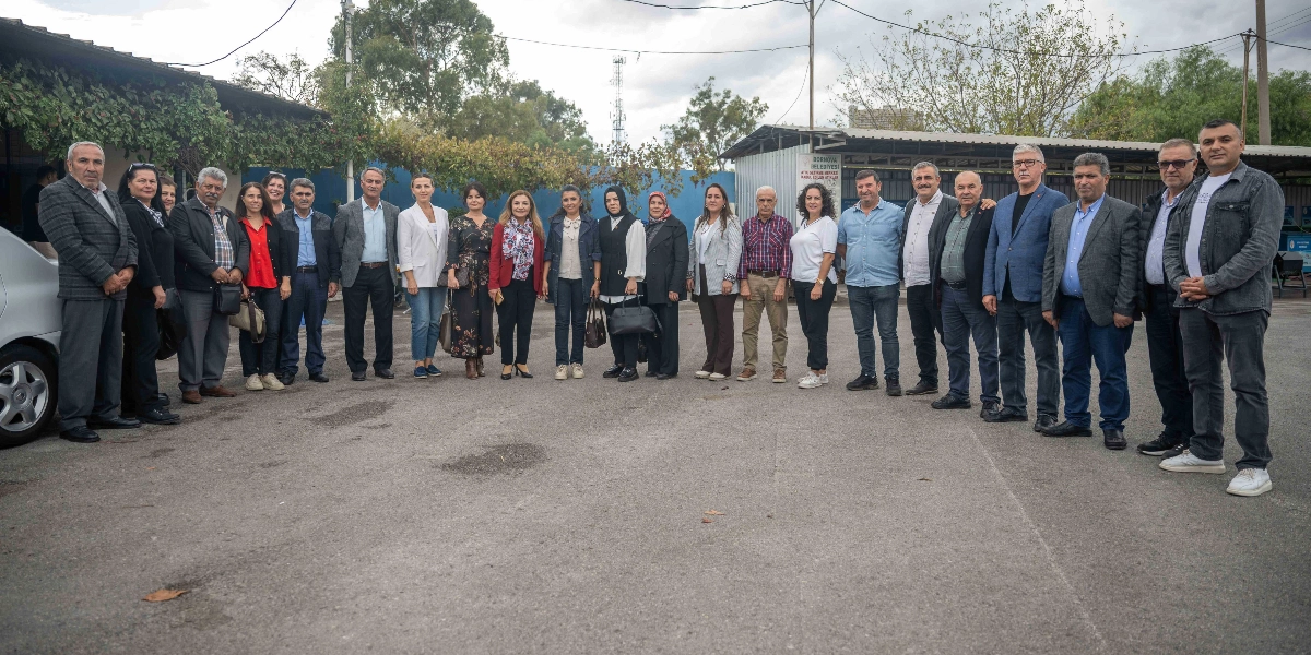 Açık havada, asfalt bir alanda yan yana dizilmiş, Bornova Belediyesi yetkilileri ve çalışanlarından oluşan geniş bir grup insan, sıfır atık projesi kapsamında bir araya gelerek poz veriyor. Arka planda Bornova Belediyesi'ne ait bir atık toplama merkezi ve ağaçlar yer alıyor.