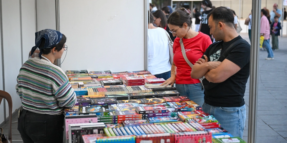 Bir kitap fuarı standında, çizgili kazaklı ve başında bandana olan bir görevli, önündeki masada dizili kitapları inceleyen kırmızı tişörtlü bir kadın ve siyah tişörtlü bir erkekle ilgileniyor. Masanın üzerinde çok sayıda farklı türde kitap sergileniyor. Arka planda ise fuar alanında gezinen diğer ziyaretçiler görülüyor.
