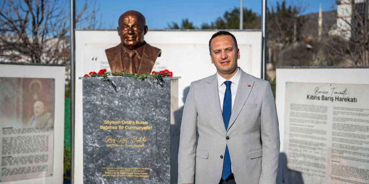 Gri takım elbiseli ve mavi kravatlı bir erkek, arka planda Rauf Denktaş’ın büstünün bulunduğu anıtın önünde duruyor. Büstün altındaki mermer kaidede 