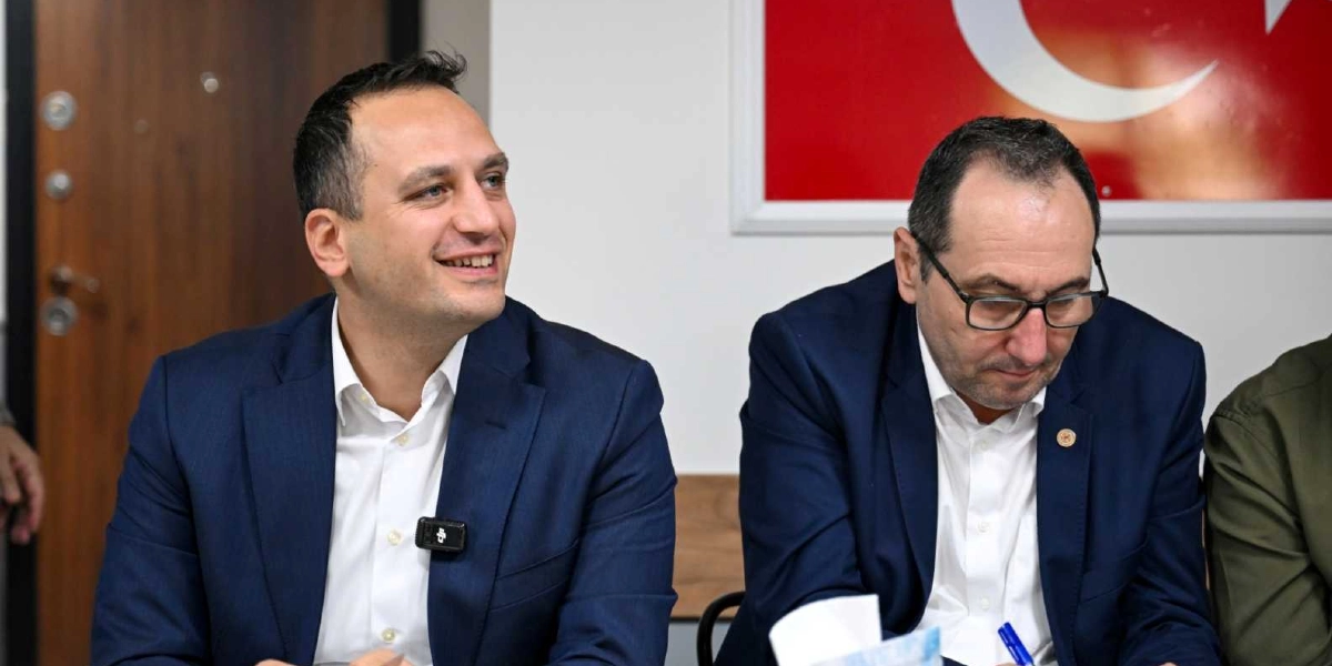 Beyaz gömlek ve lacivert ceket giymiş, gülümseyen bir erkek ile yanında oturan, gözlüklü ve lacivert ceketli başka bir erkek bir toplantı masasında yan yana oturuyor. Arka planda kısmen görünen Türk bayrağı yer alıyor.
