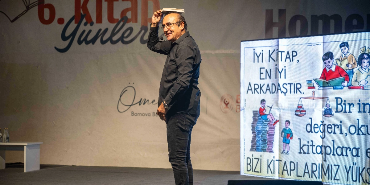 Siyah gömlek ve pantolon giymiş, gözlüklü bir erkek, sahnede başının üzerinde bir kitap dengelerken gülümsüyor. Arka planda 