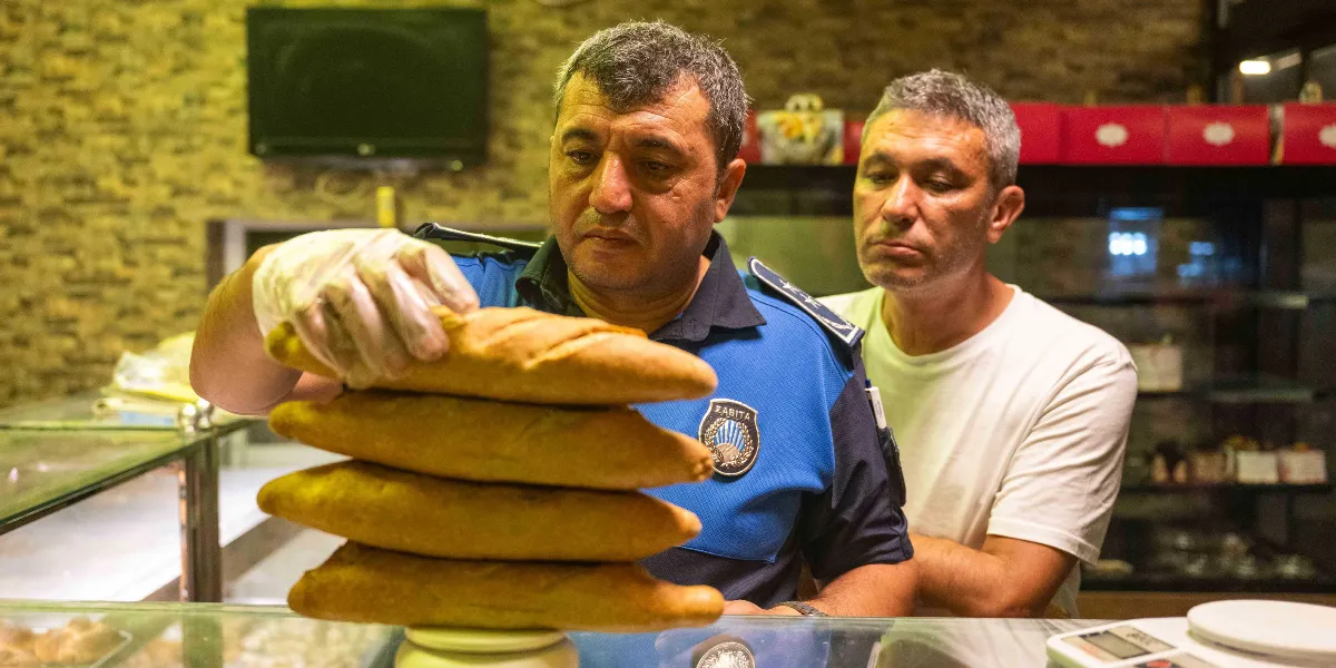 Mavi zabıta üniforması giyen bir görevli, eldivenli eliyle bir fırındaki ekmekleri kontrol ederken, yanında beyaz tişörtlü bir çalışan onu izliyor.