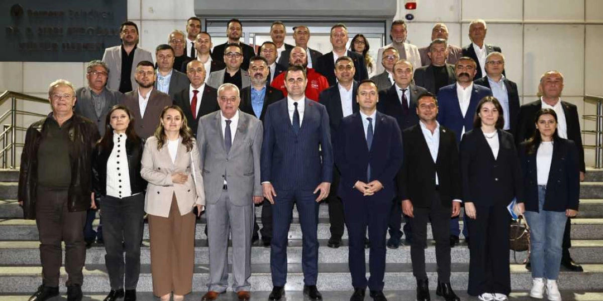 Bornova Belediyesi Dr. S. Nuri Aydın Kültür Merkezi'nin merdivenlerinde yan yana duran, çoğunluğu takım elbiseli erkekler ve kadınlardan oluşan kalabalık bir grup. Ön sırada yer alan kişiler arasında, merkezde lacivert takım elbiseli ve kravatlı bir erkek ile onun yanında yine takım elbiseli diğer siyasetçiler ve kadın yöneticiler bulunuyor. Arka sıralarda ise farklı yaş ve giyim tarzlarında, resmi kıyafetli birçok kişi yer alıyor. Grup, objektiflere gülümseyerek poz veriyor.