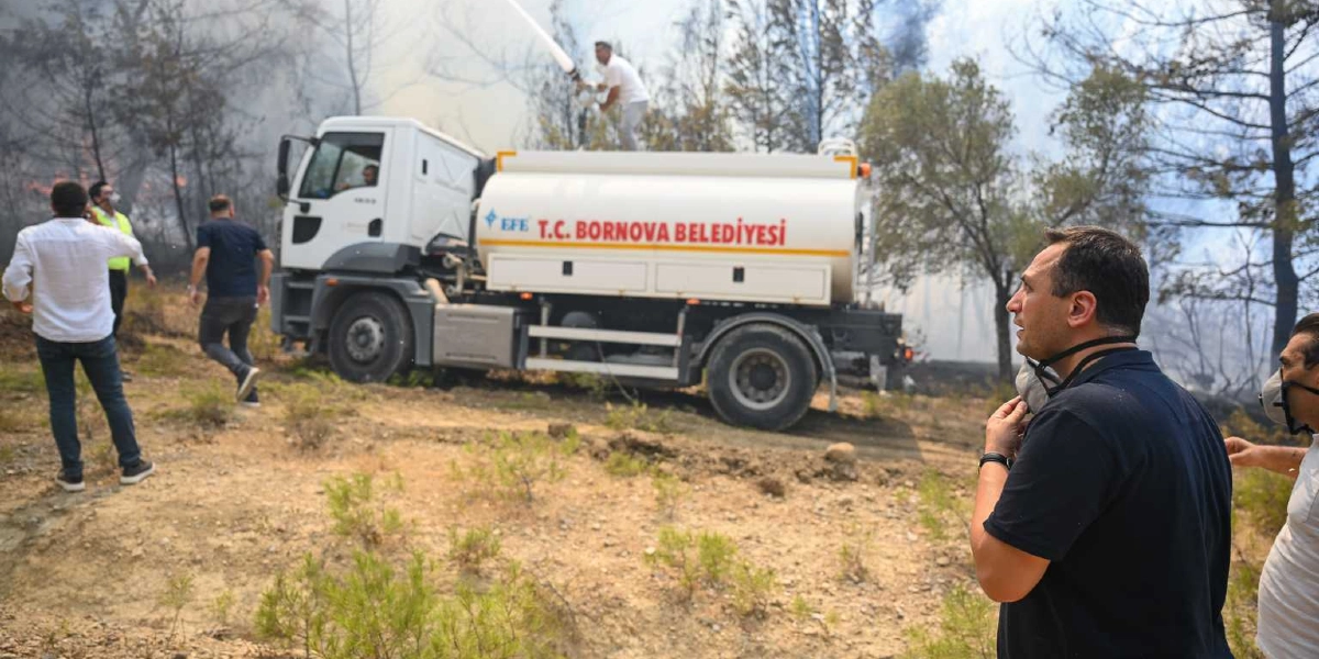 Bornova Belediyesi'ne ait beyaz bir arazözün ormanlık alandaki yangına müdahale ettiği sırada, bir görevli aracın üzerindeki hortumla su sıkarken, çevredeki yetkililer ve vatandaşlar çalışmaları izliyor. Arka planda ağaçların arasından dumanlar yükselirken, ön planda maske takan ve çalışmaları takip eden insanlar yer alıyor.