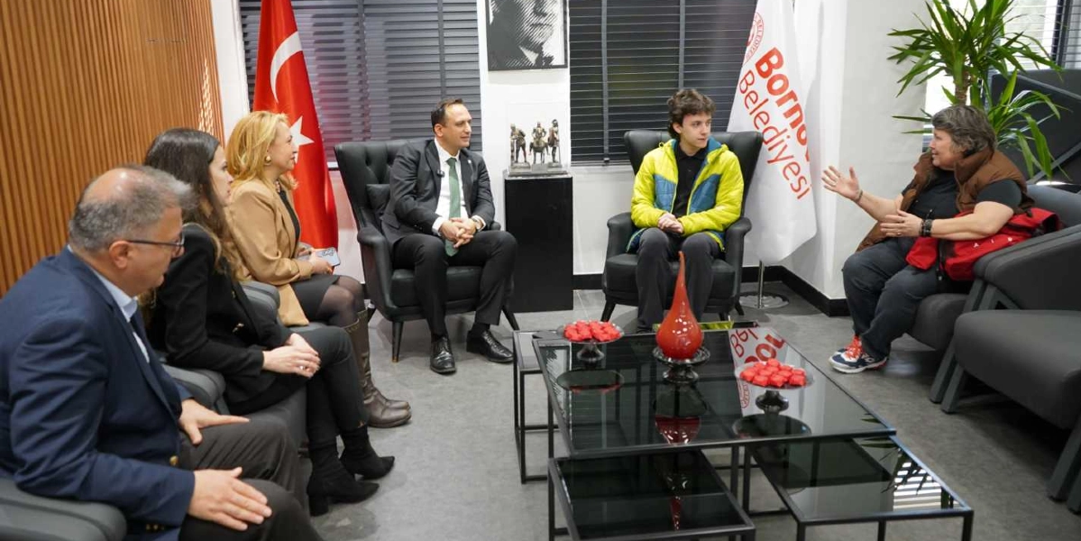 Bornova Belediye Başkanı, makam odasında bir genç ve beraberindekilerle bir araya gelerek sohbet ediyor. Odanın bir köşesinde Türk bayrağı ve Bornova Belediyesi flaması yer alırken, masanın üzerinde kırmızı süs eşyaları bulunuyor.