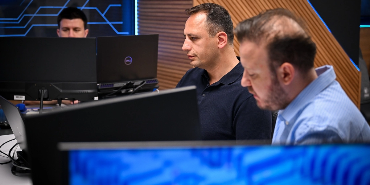 Modern bir ofis ortamında, bilgisayar ekranlarının önünde yan yana oturarak çalışan üç erkek. Ön planda, açık mavi gömlekli bir kişi ve yanında lacivert polo yaka tişörtlü bir adam dikkatle ekranlara bakıyor. Arka planda ise başka bir kişi daha bilgisayar başında çalışıyor. Çalışma alanının arka duvarında ahşap paneller ve mavi ışıklı dijital grafik desenleri bulunuyor.