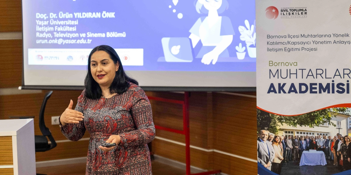 Doç. Dr. Ürün Yıldıran Önk, Bornova Muhtarlar Akademisi kapsamında bir sunum yaparken görülüyor. Arkasındaki projeksiyon perdesinde ismi, unvanı ve iletişim bilgileri yer alıyor. Sağ tarafta ise İçişleri Bakanlığı Sivil Toplumla İlişkiler Genel Müdürlüğü logolu, Bornova Muhtarlar Akademisi projesini tanıtan ve muhtarların toplu fotoğrafının bulunduğu dikey bir afiş duruyor.