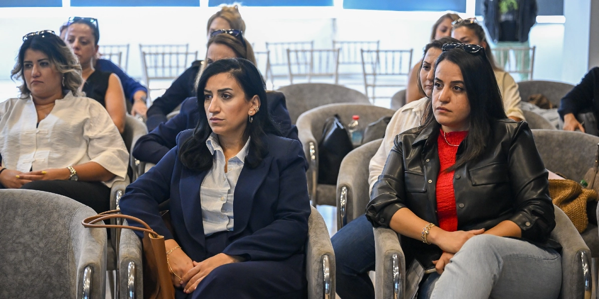 Aydınlık bir salonda, gri koltuklarda yan yana oturan ve dikkatle karşıya bakan bir grup kadın katılımcı. Ön planda, lacivert ceketli ve beyaz gömlekli bir kadın ile yanında kırmızı bluz ve siyah deri ceket giymiş başka bir kadın yer alıyor. Arka sıralarda ise farklı kıyafetler içindeki diğer katılımcılar izleyici konumunda oturuyor.