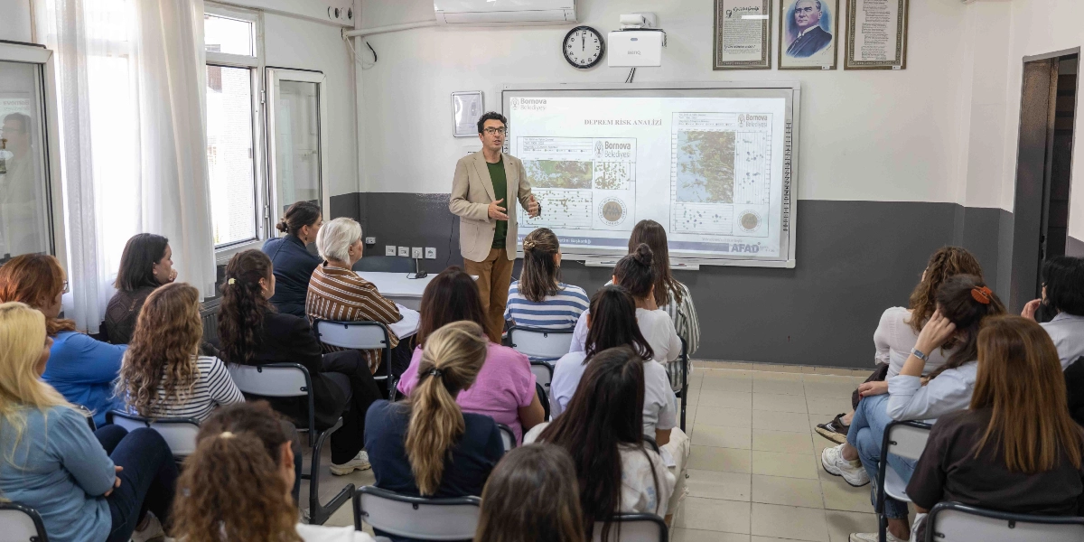 Bir eğitim sınıfında, gözlüklü bir eğitmen akıllı tahta önünde durarak katılımcılara sunum yapıyor. Akıllı tahtada Bornova Belediyesi logolu 