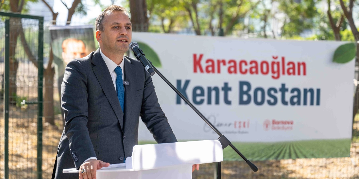 Koyu gri takım elbiseli ve mavi kravatlı bir erkek, önünde mikrofon bulunan bir kürsüde konuşma yapıyor. Arka planda üzerinde 