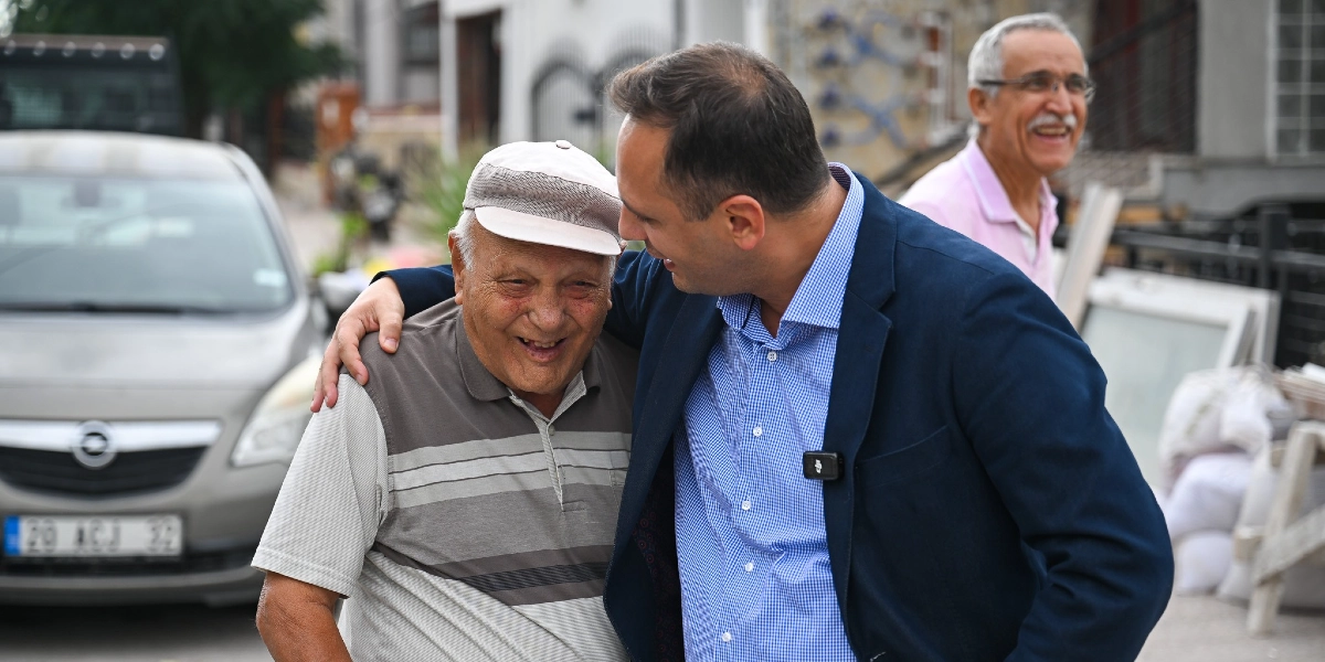 Koyu renk ceketli ve mavi kareli gömlekli bir adam, yanında duran şapkalı yaşlı bir adamın omzuna elini atarak onunla samimi bir şekilde sohbet ediyor. İkisi de gülümseyerek birbirine bakıyor. Arka planda ise açık renkli bir otomobil ve gülümseyen başka bir yaşlı adam daha bulunuyor.