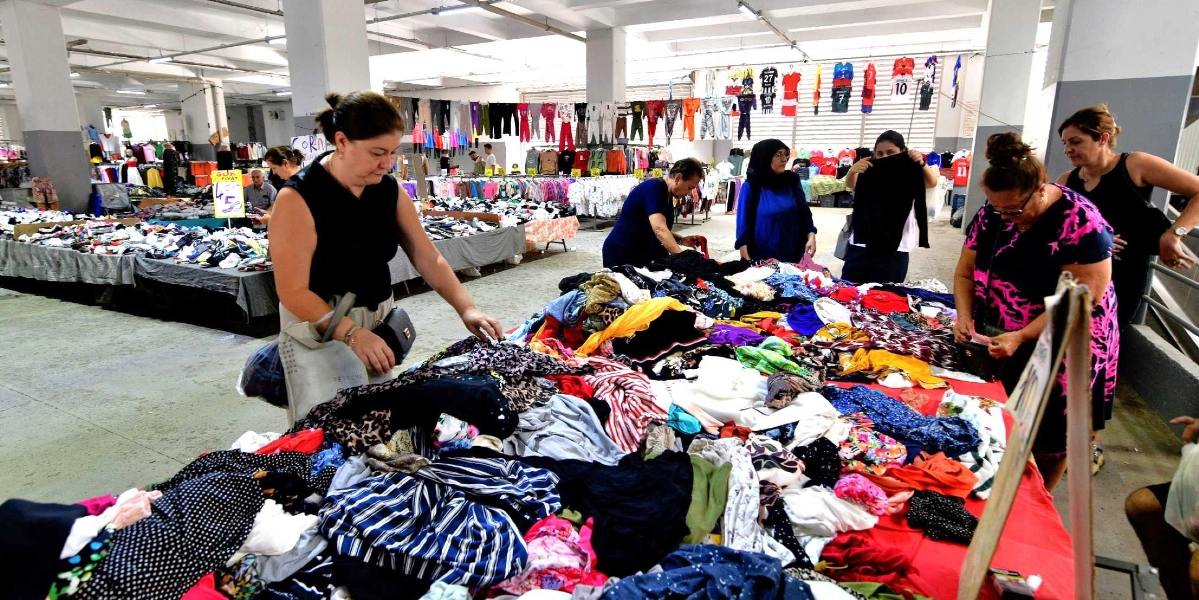 Kapalı bir pazar yerinde, üzerinde çok sayıda farklı renk ve desende kıyafetin yığılı olduğu uzun bir tezgâhın etrafında alışveriş yapan kadınlar. Arka planda askılara dizilmiş kıyafetler ve spor formaları görülüyor.