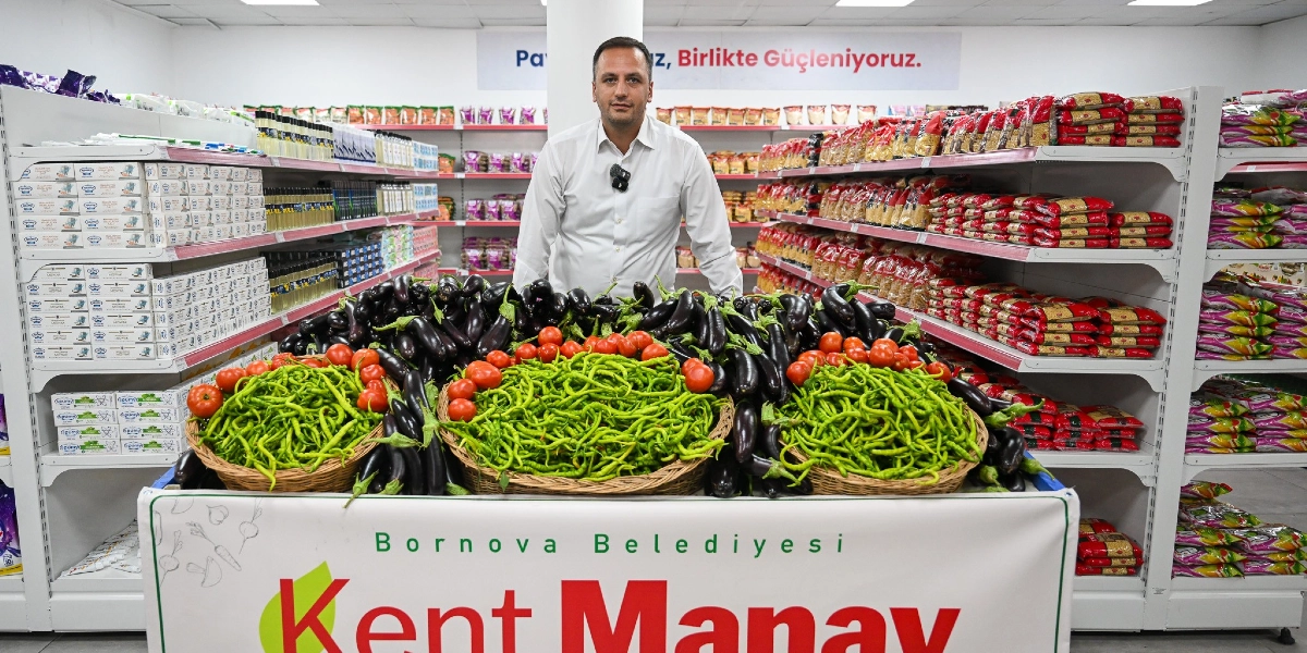 Beyaz gömlekli bir erkek, Bornova Belediyesi Kent Manavı reyonunun arkasında duruyor. Önündeki üç büyük hasır sepet yeşil biber ve domateslerle dolu, etrafı ise patlıcanlarla çevrili. Arka planda raflarda paketlenmiş gıda ürünleri dizili ve duvarda 