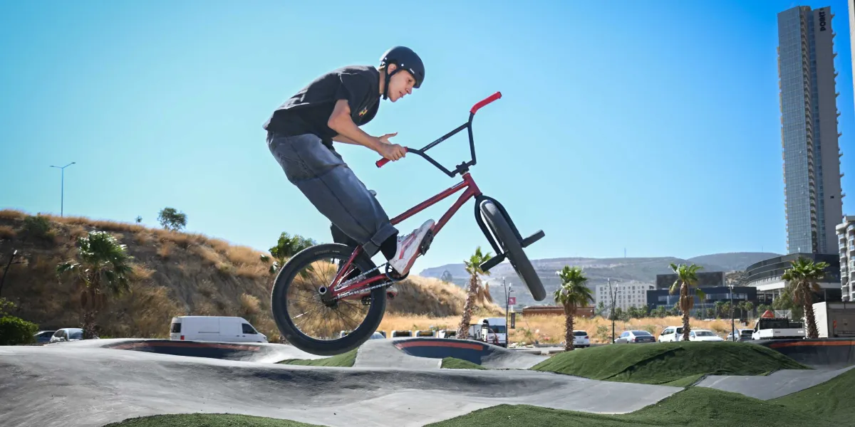 Kask takan genç bir bisikletçi, BMX bisikletiyle Bornova'daki bir parkurda havaya sıçrayarak akrobatik bir hareket sergiliyor. Arka planda palmiye ağaçları, tepelik bir arazi ve yüksek bir bina yapısı görülüyor.