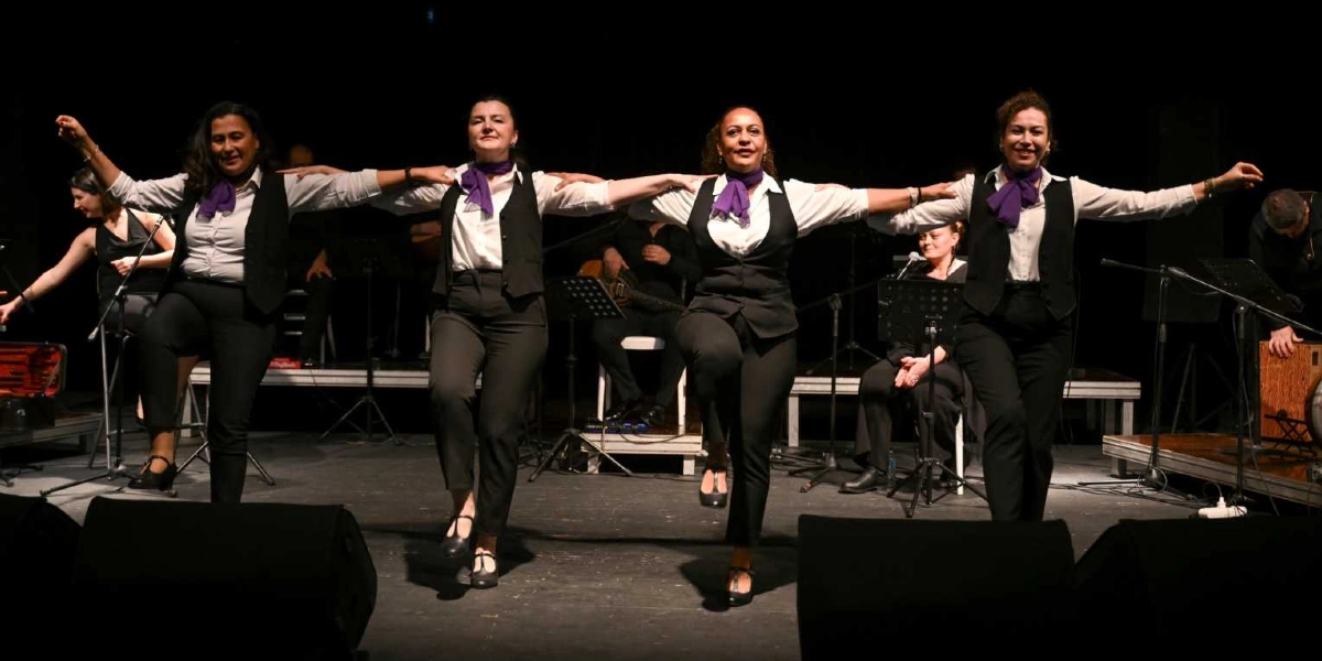 Sahne üzerinde yan yana dizilmiş, beyaz gömlek, siyah yelek ve pantolon giyen, boyunlarında mor fular bulunan dört kadın dansçı, kollarıyla birbirlerine kenetlenerek halk oyunu sergiliyor. Dansçılar bir bacaklarını havaya kaldırmış, gülümseyerek ileriye bakıyor. Arka planda ise müzik aletleriyle oturan diğer grup üyeleri ve sahne ekipmanları yer alıyor.