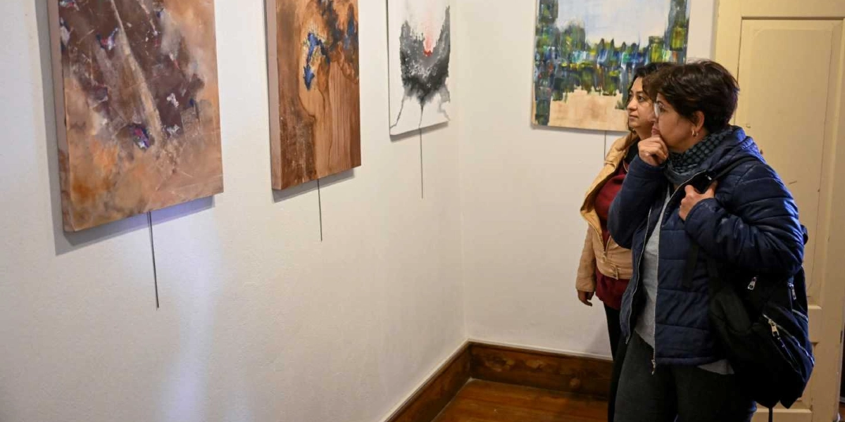 Sanat galerisinde duvarda asılı soyut tabloları inceleyen iki kadın. Önde duran kısa saçlı kadın, elini çenesine koymuş dikkatle bir esere bakarken, arkasındaki kadın da ona eşlik ediyor. Duvarlarda kahverengi tonlarının hakim olduğu dokulu çalışmalar ve beyaz zemin üzerine siyah-kırmızı detaylı bir tablo yer alıyor.