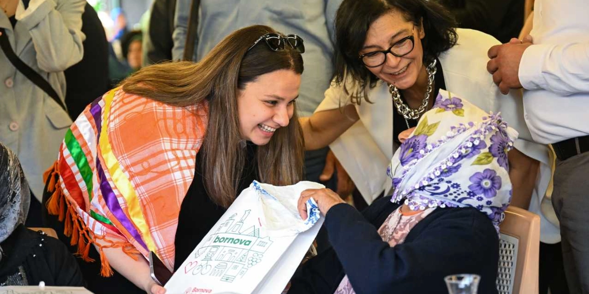Omuzlarında turuncu ve beyaz desenli bir şal bulunan genç bir kadın, üzerinde Bornova Belediyesi logosu ve çizimleri olan beyaz bir hediye çantasını, başında çiçekli bir yazma olan yaşlı bir kadına gülümseyerek uzatıyor. Arkalarında gözlüklü, beyaz ceketli bir kadın da bu anı gülümseyerek izliyor.