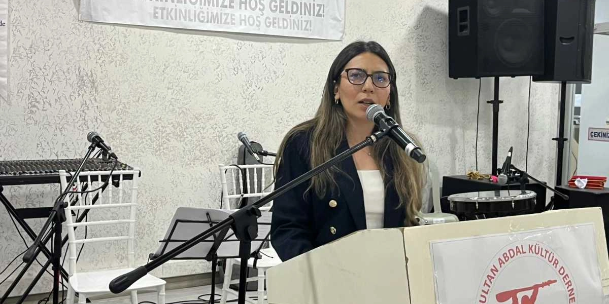 Gözlüklü, uzun kahverengi saçlı bir kadın, üzerinde Pir Sultan Abdal Kültür Derneği logosu bulunan bir kürsüde mikrofon başında konuşma yapıyor. Arka planda beyaz sandalyeler, ses sistemi ekipmanları ve üzerinde 