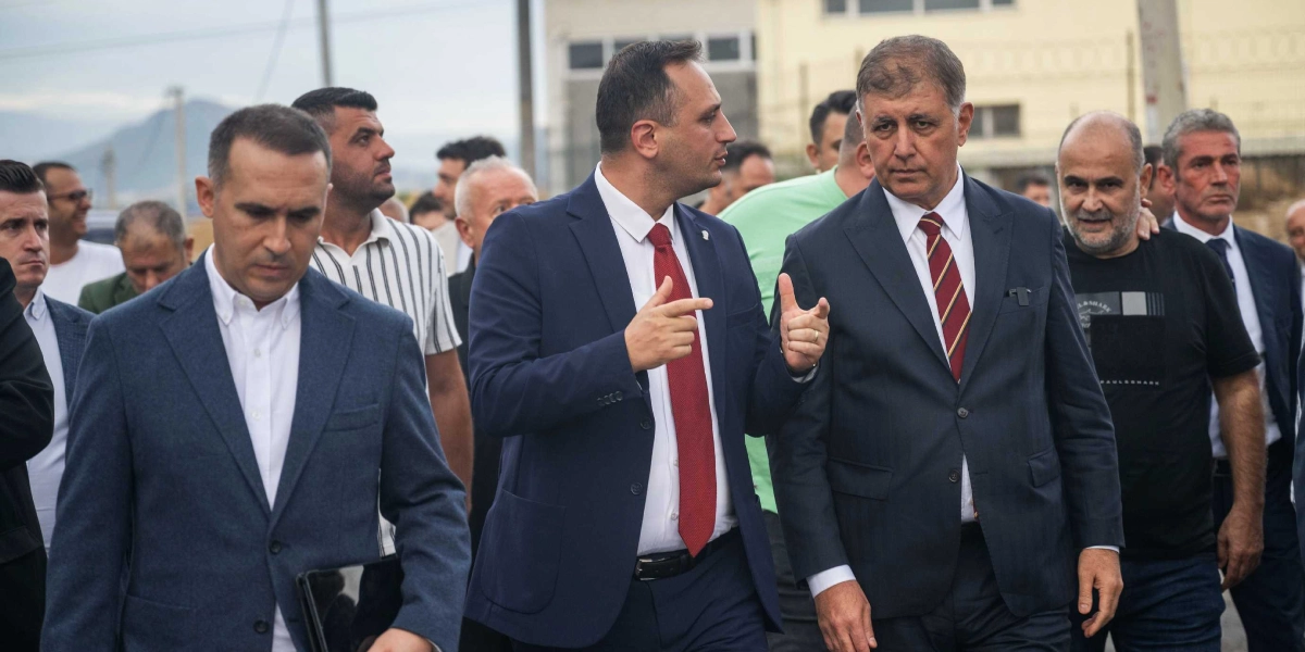 Takım elbiseli ve kravatlı iki erkek, yanlarında yürüyen kalabalık bir grupla birlikte dış mekanda ilerlerken bir konu hakkında konuşuyor. Ortadaki kişi el hareketleriyle açıklama yaparken, yanındaki kişi onu dinliyor. Arka planda ise çeşitli kıyafetler giymiş diğer erkekler yer alıyor.