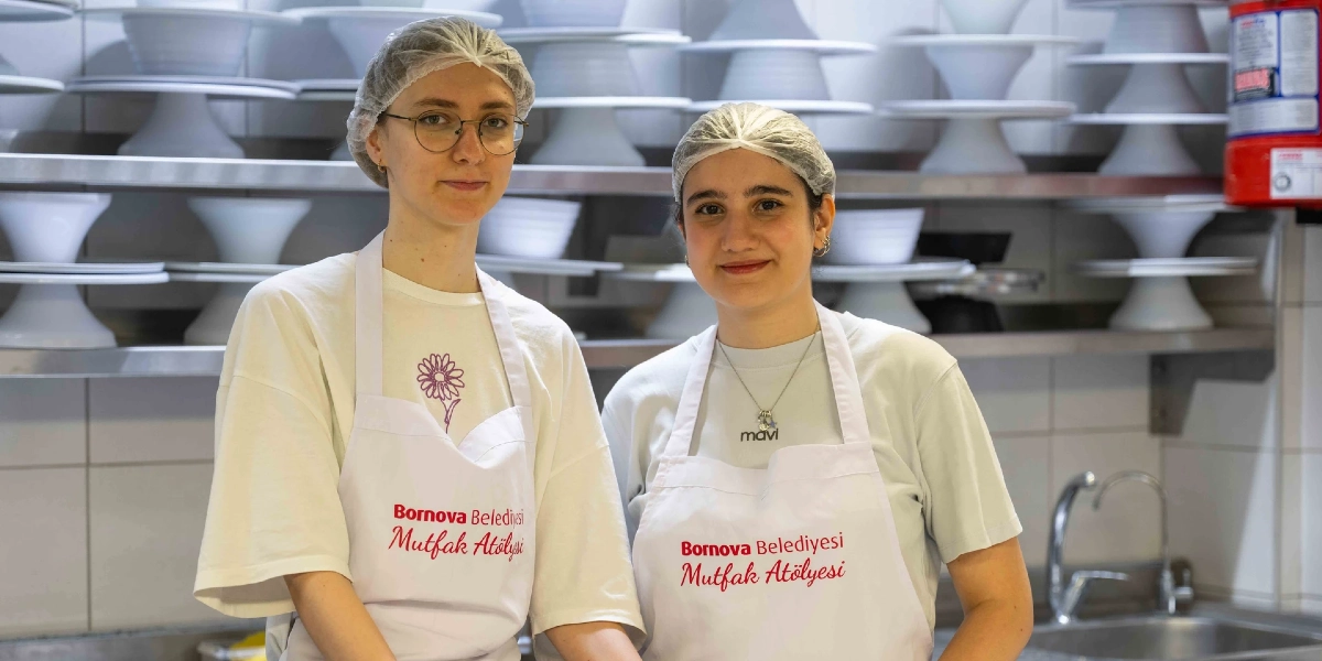 Üzerlerinde Bornova Belediyesi Mutfak Atölyesi yazılı beyaz önlükler ve saç bonesi takan iki genç kadın, arka planda raflarda dizili beyaz servis tabaklarının bulunduğu profesyonel bir mutfakta yan yana durarak gülümsüyor.