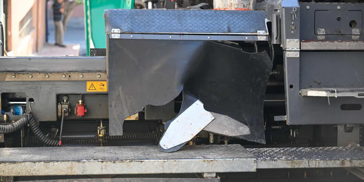 Belediyeye ait bir iş makinesinin yan tarafında, siyah plastik koruyucu panelin yırtılmış ve hasar görmüş olduğu görülüyor. Metal aksamın üzerinde sarı renkli bir uyarı etiketi ve çeşitli kablo bağlantıları bulunuyor.