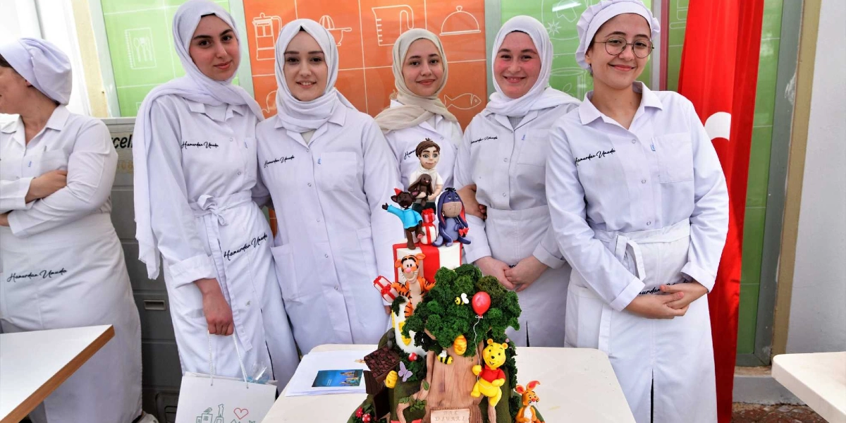 Beyaz önlük ve başörtüsü giymiş beş genç kadın, önlerinde duran Winnie the Pooh temalı, ağaç figürlü ve renkli karakterlerle süslenmiş büyük bir pastanın arkasında yan yana duruyor. Kadınların önlüklerinin üzerinde 