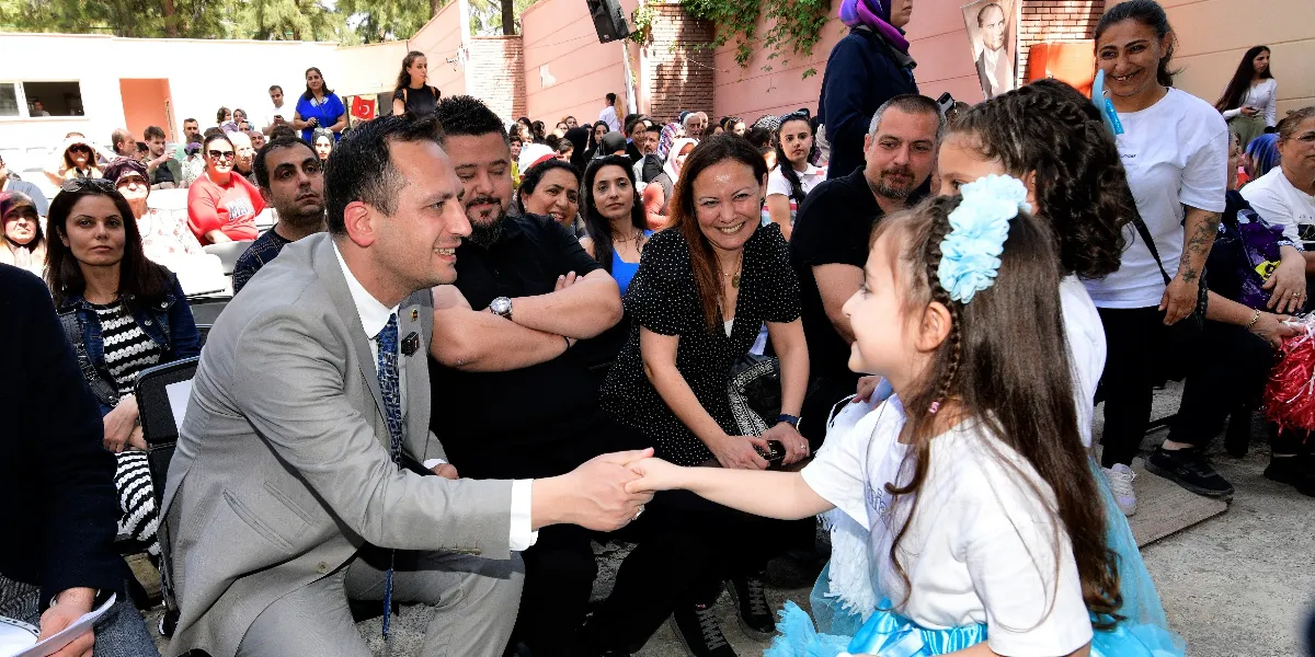 Gri takım elbiseli bir adam, açık havada düzenlenen bir etkinlikte, saçında mavi çiçekli bir toka bulunan ve mavi tüllü bir etek giyen küçük bir kız çocuğunun elini tutarak onunla gülümseyerek sohbet ediyor. Çevredeki sandalyelerde oturan yetişkinler ve çocuklar, bu anı izleyerek gülümseyen yüzlerle olaya eşlik ediyor. Arka planda ise ağaçlar ve duvarda asılı bir Atatürk portresi görülüyor.