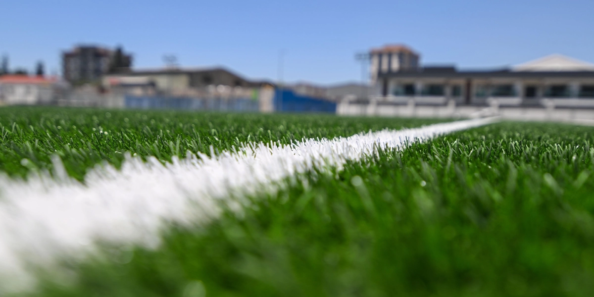 Güneşli bir günde, suni çimle kaplı bir futbol sahasının zeminine yakın çekim odaklanılmış. Sahayı boydan boya geçen beyaz bir çizgi, net bir şekilde ön planda yer alırken, arka planda bulanık bir şekilde görünen binalar ve açık mavi bir gökyüzü bulunuyor.