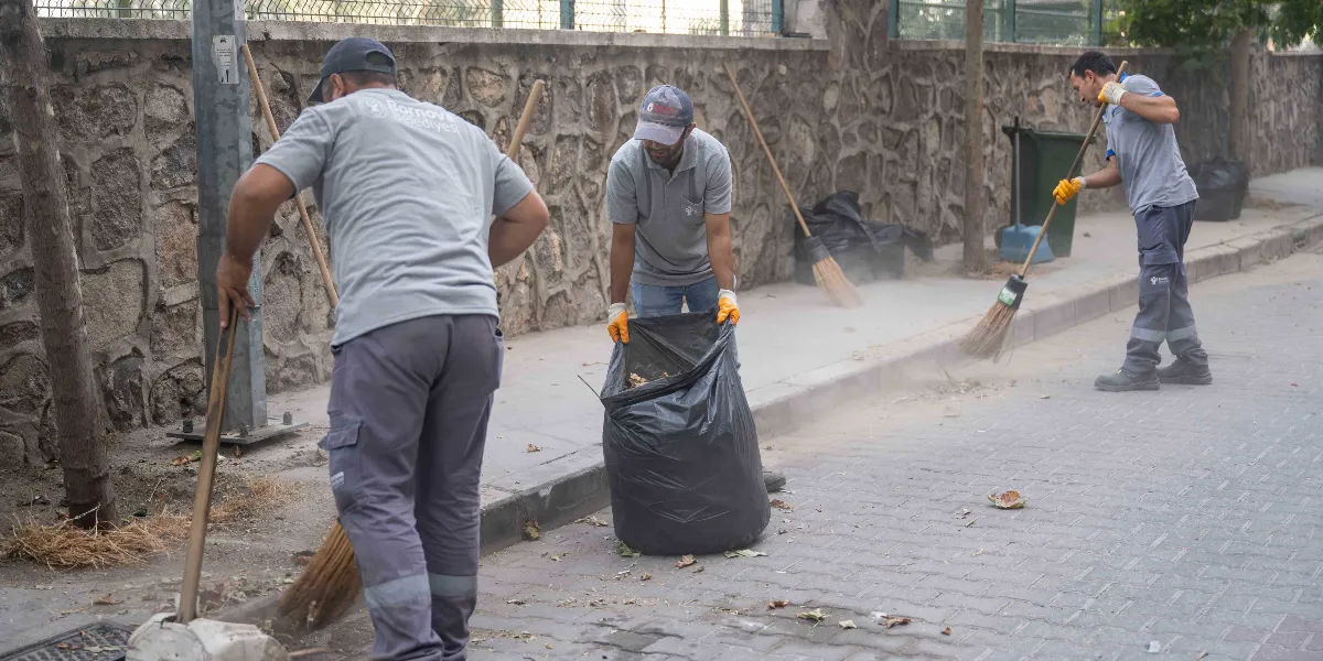 Bornova Belediyesi üniformalı üç temizlik işçisi, taş duvarlı bir sokakta süpürge ve çöp poşetleriyle temizlik yapıyor. İşçilerden biri büyük bir çöp poşetini açık tutarken, diğer ikisi ellerindeki süpürgelerle kaldırım kenarındaki toz ve yaprakları temizliyor.