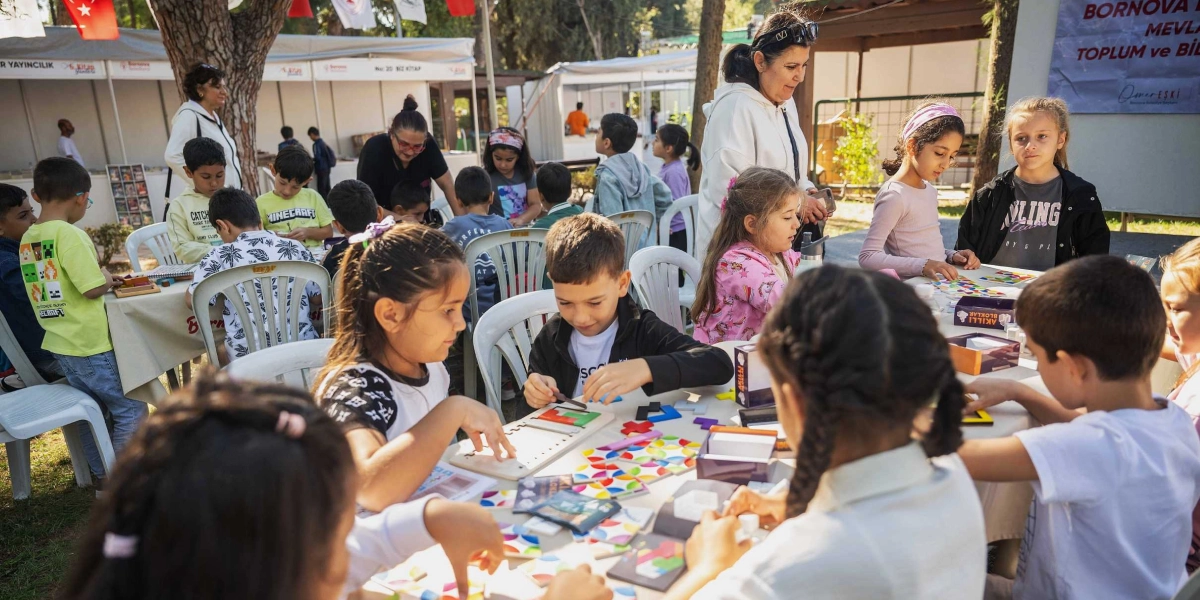 Açık havada, ağaçların altında kurulan stantların önünde, beyaz plastik masaların etrafında toplanmış çocuklar zeka oyunları ve yapbozlarla vakit geçiriyor. Çocuklar dikkatle oyun parçalarını birleştirirken, yanlarında onlara eşlik eden yetişkinler bulunuyor. Arka planda Bornova Belediyesi'ne ait stantlar ve üzerinde 