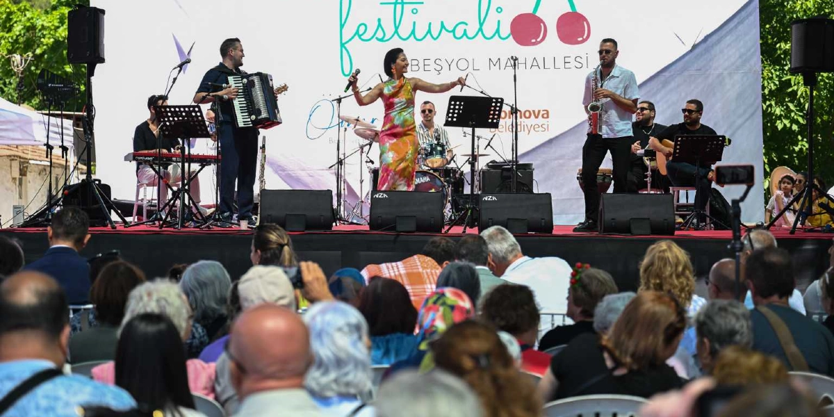 Bornova Beşyol Mahallesi'nde düzenlenen kiraz festivalinde, sahnede canlı müzik yapan bir grup ve onları izleyen kalabalık bir kitle görülüyor. Sahnedeki müzisyenler arasında akordeon çalan bir kişi, mikrofonla şarkı söyleyen renkli elbiseli bir kadın, saksafon çalan bir erkek, baterist ve gitarist yer alıyor. Arka planda üzerinde kiraz figürleri ve festival yazısı bulunan büyük bir sahne dekoru bulunuyor. İzleyiciler ise sahnenin önündeki sandalyelerde oturarak konseri takip ediyor.