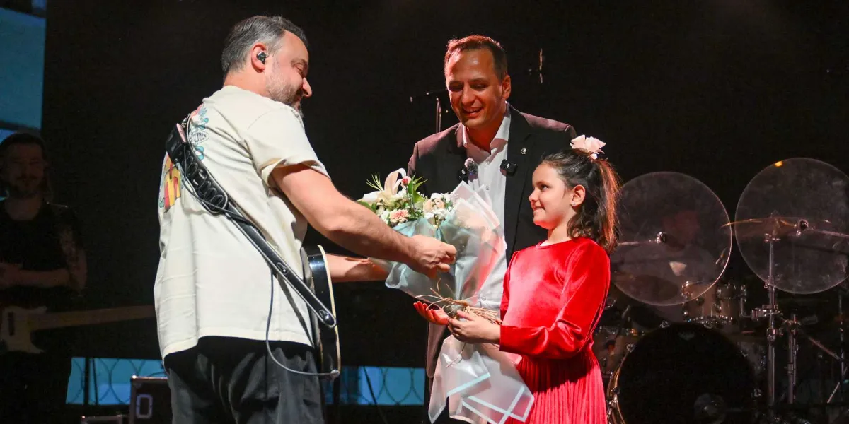 Sahne üzerinde, sırtı dönük duran ve omzunda gitar askısı bulunan beyaz tişörtlü bir müzisyen, kırmızı elbiseli küçük bir kız çocuğuna çiçek buketi veriyor. Müzisyenin yanında, siyah takım elbiseli bir adam gülümseyerek bu anı izliyor. Arka planda ise bir bateri seti ve diğer müzisyenler seçiliyor.
