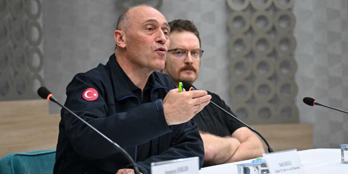 Koyu renkli, üzerinde Türk bayrağı arması bulunan bir ceket giyen orta yaşlı bir erkek, bir toplantıda konuşma yapıyor ve elindeki kalemle bir noktayı işaret ediyor. Arka planda ise gözlüklü, bıyıklı ve siyah tişörtlü bir başka erkek dinleyici konumunda oturuyor. Önlerinde mikrofonlar ve isimliklerin bulunduğu beyaz örtülü bir masa yer alıyor.