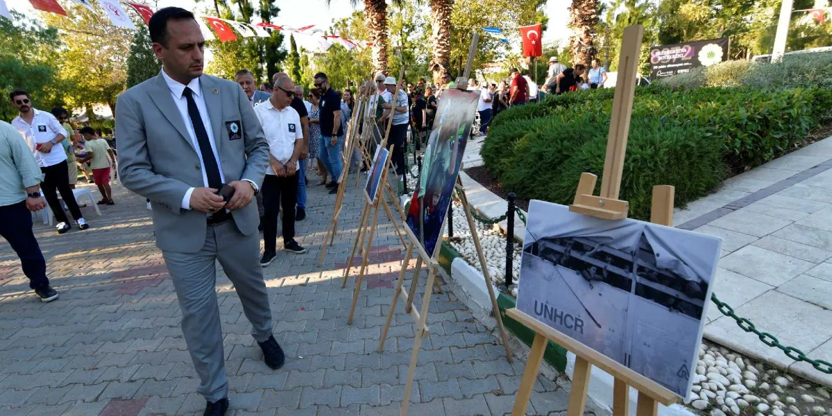 Açık havada düzenlenen Srebrenica anma sergisinde, gri takım elbiseli bir adam şövale üzerindeki fotoğrafları inceliyor. Park içindeki yürüyüş yolunda yan yana dizilmiş şövalelerde, Srebrenica soykırımına dair siyah beyaz ve renkli fotoğraflar yer alıyor. Ön plandaki şövalede üzerinde 